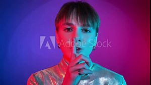 Portrait young adult woman saying hush be quiet with finger on lips shh gesture in neon illumination indoors. Studio shot in art colorful lighting. Female person short hair wearing sparkling clothing