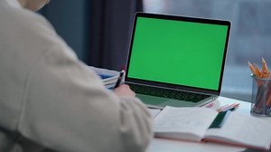 Woman Uses Laptop with Green Mock-up Screen. Girl Student Taking Notes on Notebook from Laptop at Online Lesson