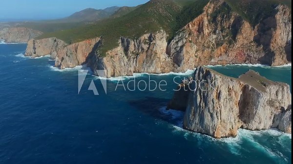 Aerial pull outfootage of Pan di Zucchero, the iconic sea stack near Masua, Sardinia. Towering limestone cliffs rise from deep blue waters, backed by green hills under clear Mediterranean skies.