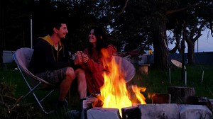 Couple in love is sitting by the outdoor campfire in the courtyard of the house on camping chairs, a romantic evening. a man and a woman warm their hands by the fire, drink tea