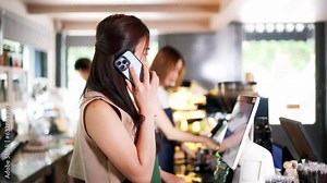 Asian coffee shop female employee accepts a pre-order on a mobile phone call while using a tablet computer in a cafe. Woman waiter talking on the cellphone. Home food delivery concept.