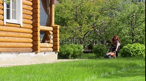 Woman afar starting lawn mower on green lawn in backyard near wooden country house on summer sunny day. Landlady cleaning up territory, growing plants background. Maintenance of home garden.