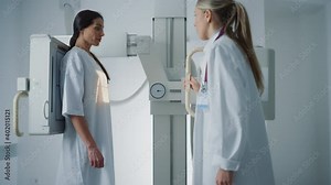 Hospital Radiology Room: Beautiful Multiethnic Woman in Medical Gown Standing Next to X-Ray Machine while Female Doctor Adjusts it. Healthy Patient Undergoes Routine Scanning with the Nurse's Help.