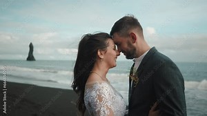 Loving smiling bride and groom with closed eyes standing face to face on beach with black sand. Blue sky, sharp rocks and hissing sea in the background. High quality 4k footage