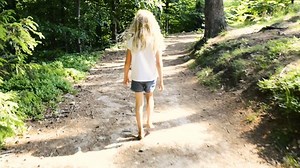 A child girl walking barefoot on a forest mountain road in bright summer light. Handheld smooth footage