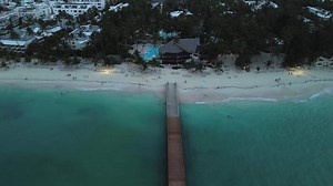 The camera gracefully rises to capture a cinematic aerial view of a pier, a tourist beach, and a hotel along the Caribbean coast at dusk, illuminated by cool lighting