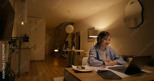 A woman in casual clothes finishes a video chat on a laptop sitting at a table at home. She takes off her headphones and closes her laptop