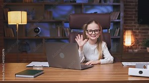 Front view of cheerful friendly little girl smiling and waving hand on camera. Positive child in formal attire posing at long office table with personal wireless computer. Greeting concept.