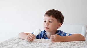 Handsome boy eating popsicle on a stick. He bites off the ice cream with pleasure. Close-up. Child eat in the kitchen or at the dining room table. Tasty dessert. Slow motion. High quality 4k footage.