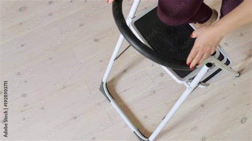 White step ladder with a black handle. The ladder is on a wooden floor. There is a person standing behind the ladder