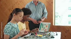 Teacher standing while explain coding and robotic construction. Diverse teenager fixing mother board and using electronic tool while looking at laptop display programming code screen. Edification