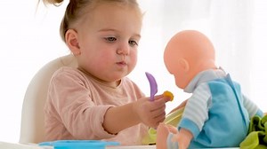 Funny little girl puffing cheeks and giving cereals to baby doll while having breakfast at home in morning