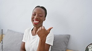 Radiant african american woman relaxing on her comfy bed, confidently pointing to the side with a thumbs-up, expressing cheeriness, and a beautiful smile in a cozy room