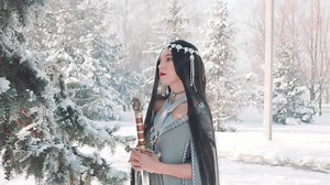 woman fantasy warrior holding royal elven sword in hand. long black hair, red lips make-up posing for camera, elf princess with blade sharp weapon. Creative dress gray cloak in winter forest pine tree