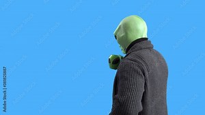 Man in alien costume, warm knitted jacket, with funny themed mug in his hand pretends to wipe the window with sleeve and drink hot beverage against a blue chroma key. Preparation for Halloween party.