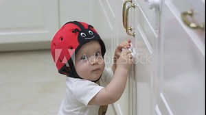 Childproofing a house, baby wearing safety helmet exploring kitchen cupboards, curious toddler trying to open a cabinet door with installed child safety lock. High quality 4k footage