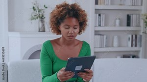 Young concentrated positive African American woman with tablet computer watches series or internet podcast in gadget and smiles after seeing funny moment sits on sofa in living room.