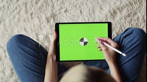 Woman sits in lotus position and draws with stylus on electronic tablet with chromakey, green screen, top view. Mockup. Concept of digital creativity