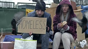 Two dirty and poorly dressed homeless people, a man and a woman, sit by a pile of rubbish with a handwritten HOMELESS poster. Couple was frightened by a flock of pigeons.