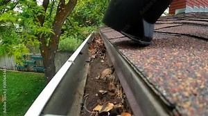 A leaf blower is seen blowing leaves and debris out of a rain gutter on a roof. A green yard with trees is seen in the background.