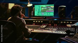 Audio technician uses mixing and mastering techniques in studio to edit and process tracks, working on console with moving faders and colored meters. Sound designer removing white noise. Camera B.