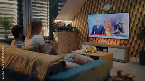 Couple sitting on couch in living room together, talking, watching TV news about war in Ukraine and Russian invasion. Man and woman spending time after work at home. TV live broadcasting.