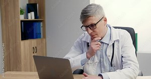 Serious man doctor sitting at workplace in hospital working on computer, looking thoughtful. Professional male physician studying patient's diagnosis.