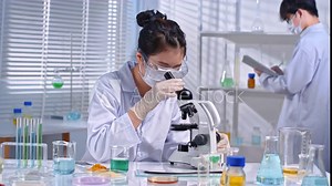 In a modern lab setting with natural light, a scientist holds a strand of hair under a microscope to observe, while another person in the back observes vials of solution and takes notes.