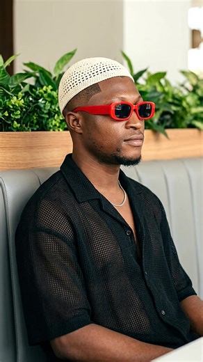 #creatorsearchinsights ai realistic photo app. copy and paste prompt. Use my uploaded image as the subject reference. Create a high-end professional portrait of a stylish Black man seated in a modern indoor lounge. He is sitting on a grey upholstered booth seat, positioned slightly sideways, with his body facing forward but his head turned to the right, looking off-frame with a calm, confident expression. He is wearing a cream/white crocheted skull cap, bold red rectangular sunglasses, a black o