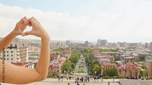 Young female tourist making heart shape with hands on famous cascade complex viewpoint with cityscape view in Yerevan, Armenia capital. Traveler having fun great vacation