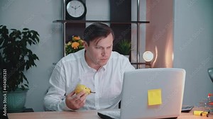 A man eats a pear in his workplace and works in front of a computer. A businessman eats in front of a computer and at an office table.