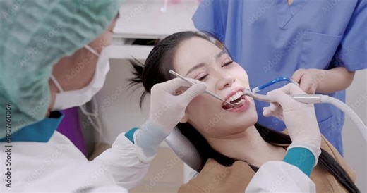 Young asian adult undergoing dental hygiene procedure while dentist operates scaler and assistant supports with suction tool during cleaning session in modern dental clinic