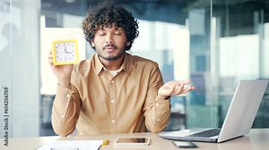 Young handsome employee holds a watch in his hands and shows looking at camera while sitting at desk at workplace in modern office. Worried man says that there is little time left to meet the deadline