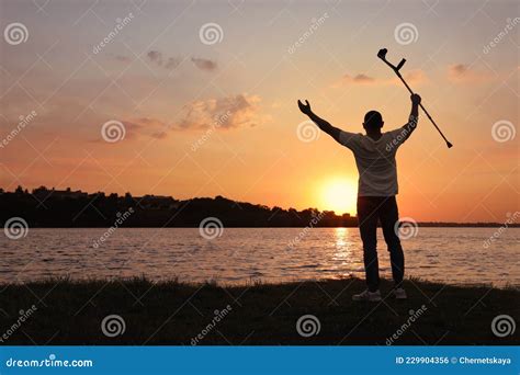Man Raising Elbow Crutch Up To Sky Near River At Sunset Healing