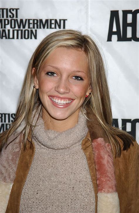 A Woman With Long Blonde Hair Smiles At The Camera While Wearing A Brown Jacket And Scarf