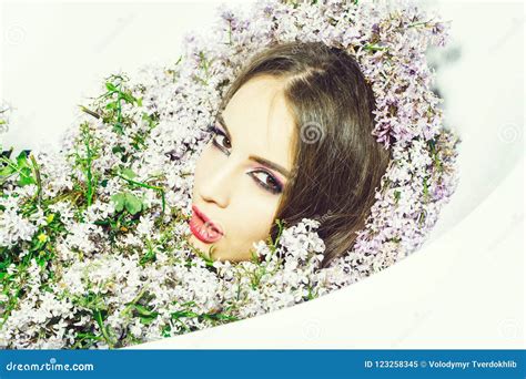 Attractive Girl Naked With Dark Hair And Evening Makeup Enjoys Bath With Lilac Petals On White