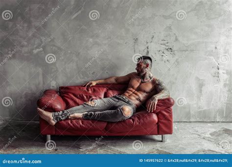 Fashion Portrait Of Naked Male Model With Tattoo And A Black Beard Lying In Hot Pose On Red