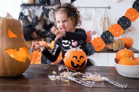 Cute Girl In Costume Of Witch With Pumpkin At Home In Kitchen Having Fun Celebrating Halloween