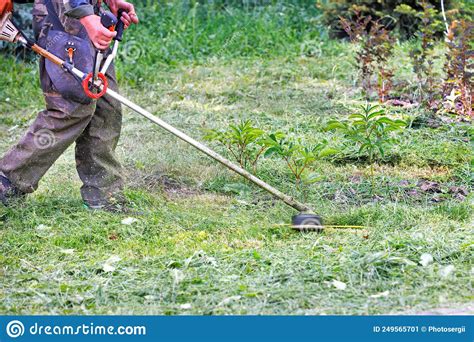A Utility Worker Mows Grass With A Petrol Trimmer Against A Green Lawn