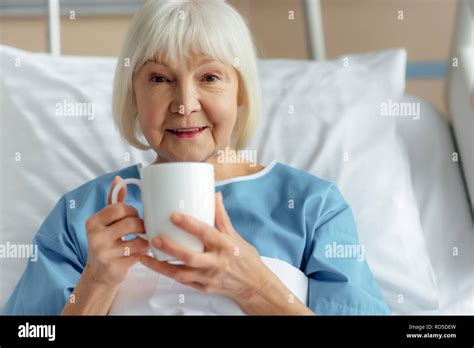 Smiling Senior Woman Lying In Bed Looking At Camera And Drinking Tea