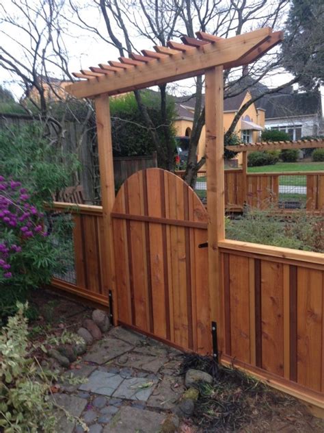 Front gate pergola boasting carriage scones entering into a long pathway aligned with palm trees in a gray pergola attached to a house finished with seeded glass carriage lanterns hanging over gray. Cedar gate and pergola | Deck Masters, LLC