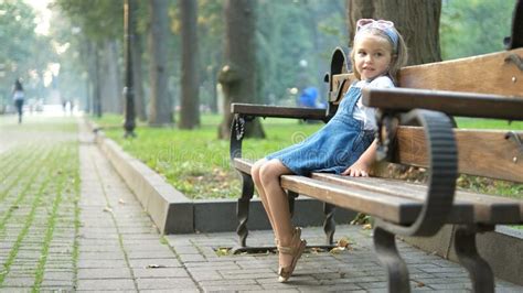 Small Happy Child Girl Sitting On A Bench Resting In Summer Park Stock