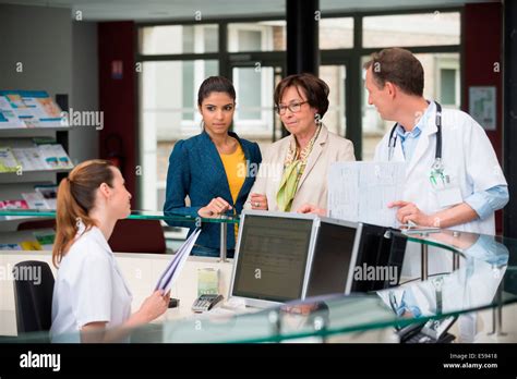 Patients Discussing With Receptionist At Hospital Reception Desk Stock