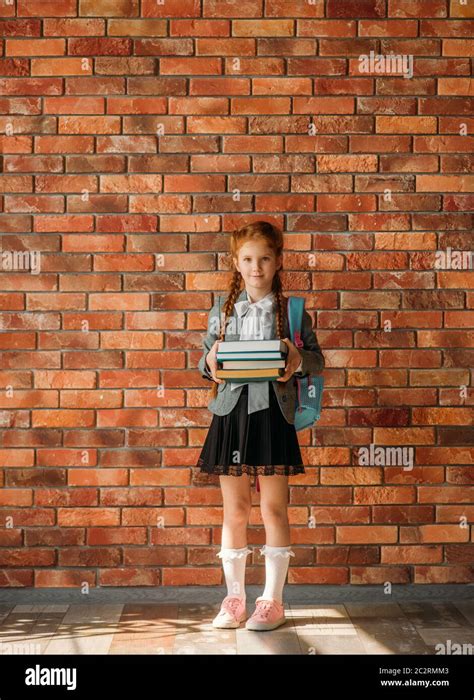 Cute Schoolgirl With Schoolbag Holds Stack Of Textbooks Brick Wall On