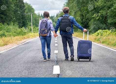 Couple Tourists With A Travel Bag On Wheels Walk Away Along The Narrow