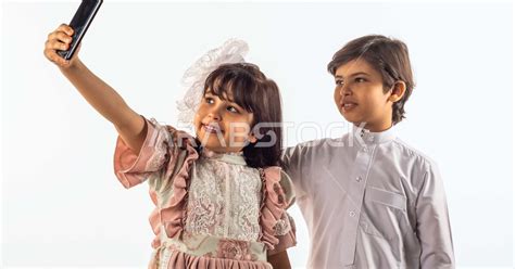 خلفية بيضاء لصبي وفتاه صغيران سعوديان مبتسمان ، تمسك الفتاه الجوال بيدها يقومان بإلتقاط صور