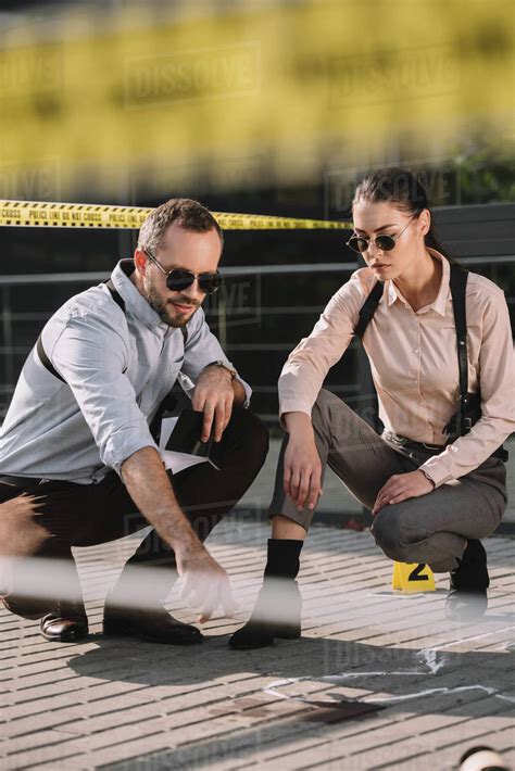 Male And Female Detectives Sitting And Looking At Chalk Line At Crime