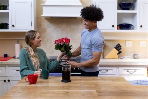 Biracial Young Husband Giving Red Roses To Happy Wife Having Coffee At Table In Kitchen Stock