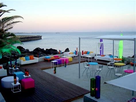 An Outdoor Seating Area Next To The Ocean At Dusk With Colorful Chairs And Tables On It