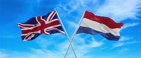 flags of great britain and netherlands waving in the wind on flagpoles against sky with clouds